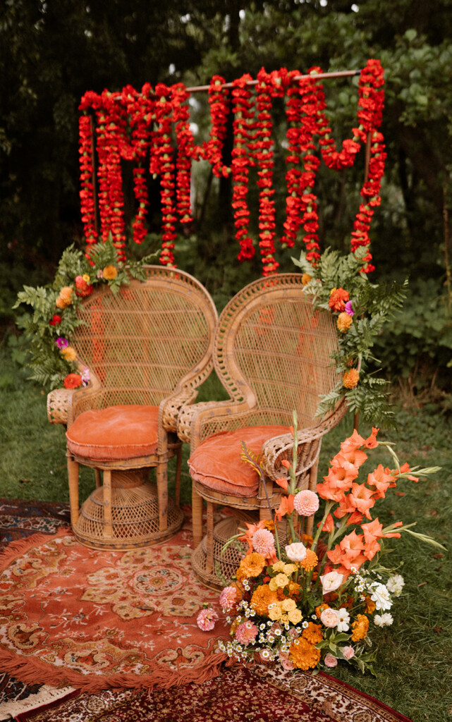 Twee pauwenstoelen met oranjerode Bruiloft decoratie eromheen. Er hangen slingers met oranjerode bloemen en er ligt een vintage kleed onder de stoelen